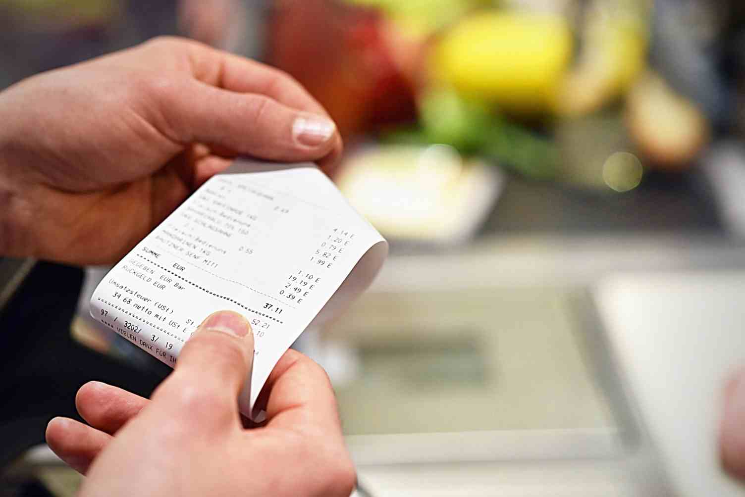 foco nas mãos de uma pessoa que está segurando uma nota fiscal de supermercado, o caixa e alguns produtos aparecem desfocados em segundo plano
