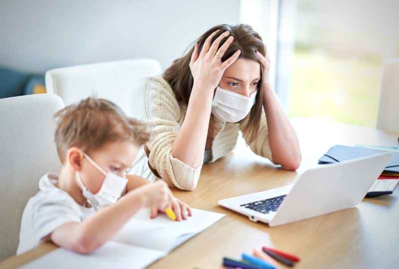 uma mãe de máscara observa o computador de maneira apreensiva enquanto um menino, também de máscara, faz lição ao seu lado na mesa