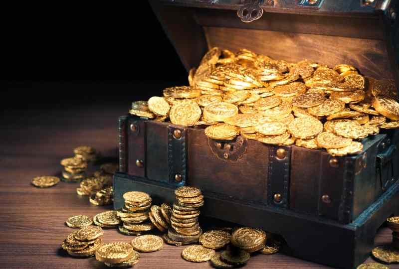 baú aberto cheio de moedas douradas dentro e algumas fora remetem à ideia de um tesouro