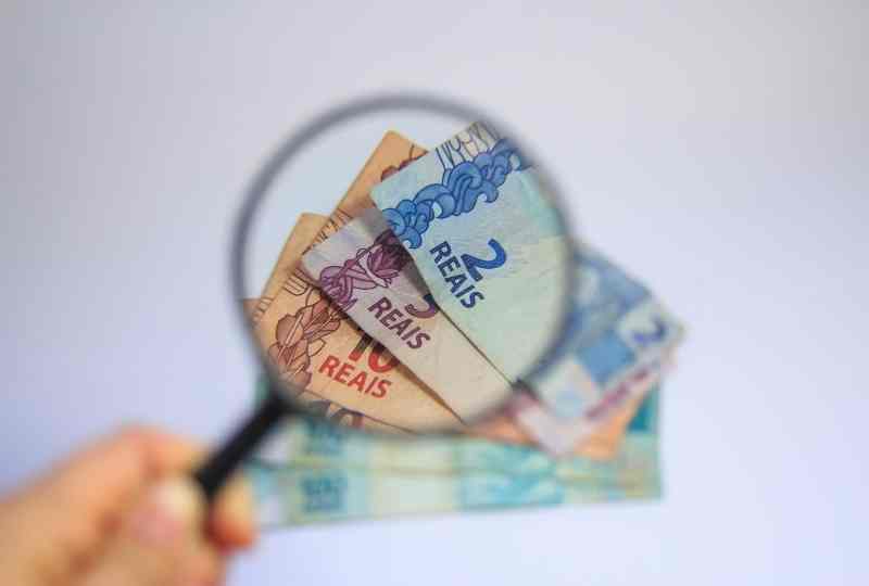 imagem mostra uma lupa focando em algumas notas de dinheiro